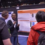 Chinna Adaikkan stands before a research poster and talks to two onlookers.