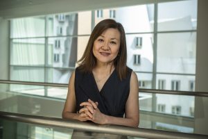 Li-Huei Tsai leans on a railing and smiles. A large window is in the background